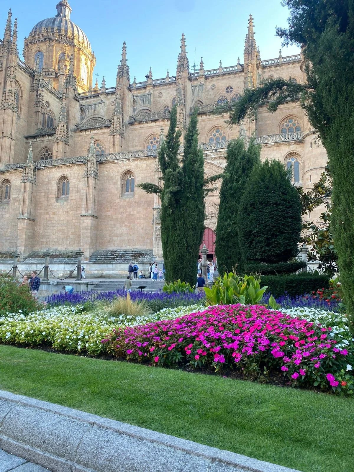 View of a historic cathedral with intricate architecture, tall spires, and a domed roof, surrounded by vibrant flower beds and tall green cypress trees, with visitors walking in the area.