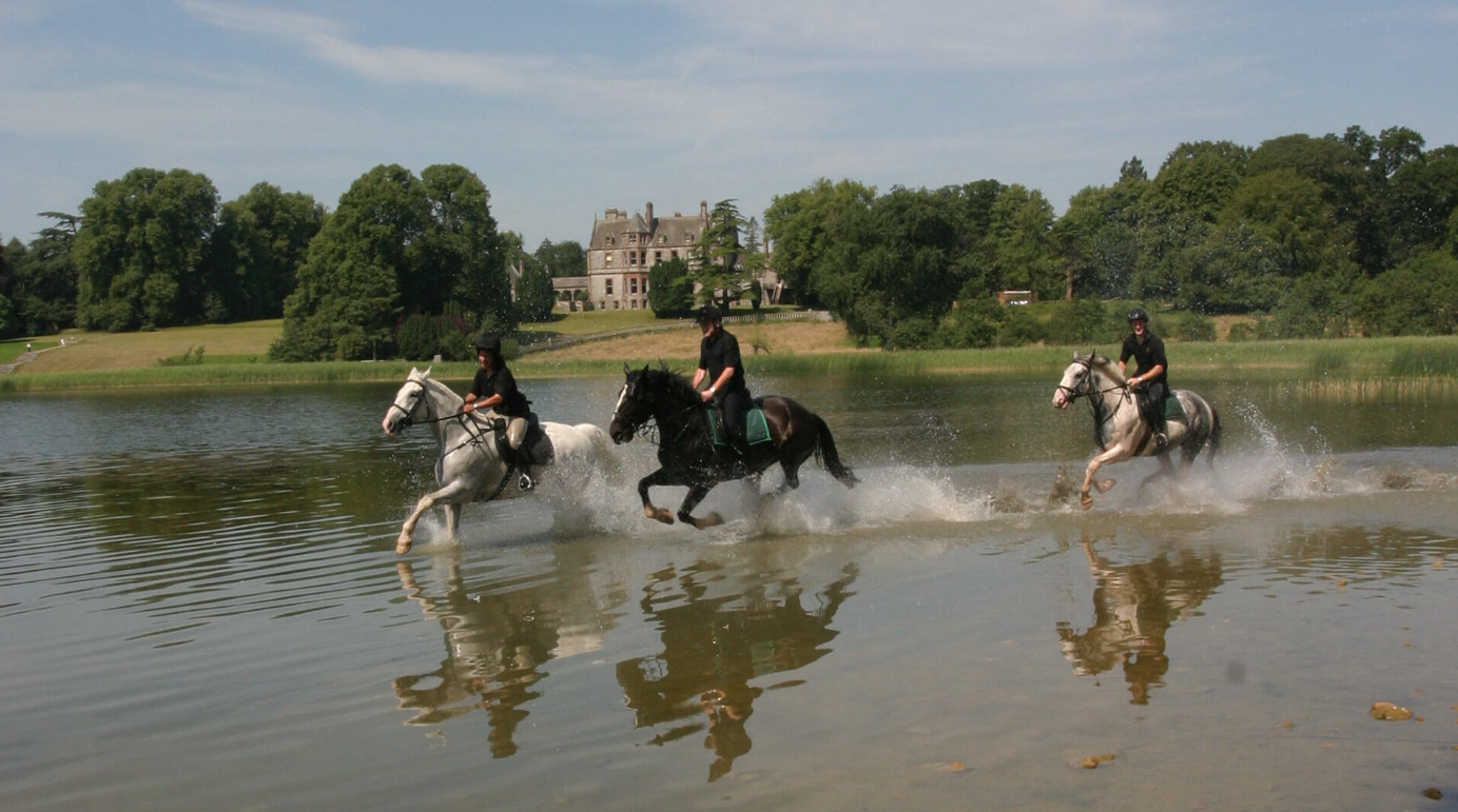 Three people riding horses through shallow water in a park-like setting with a large mansion in the background.