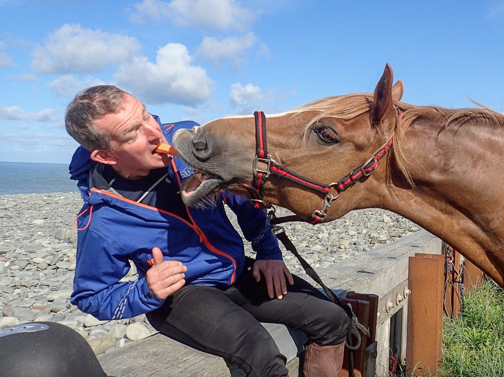 A man in a blue jacket sits on a dock by the rocky shoreline, playfully feeding a horse near the water under a partly cloudy sky.