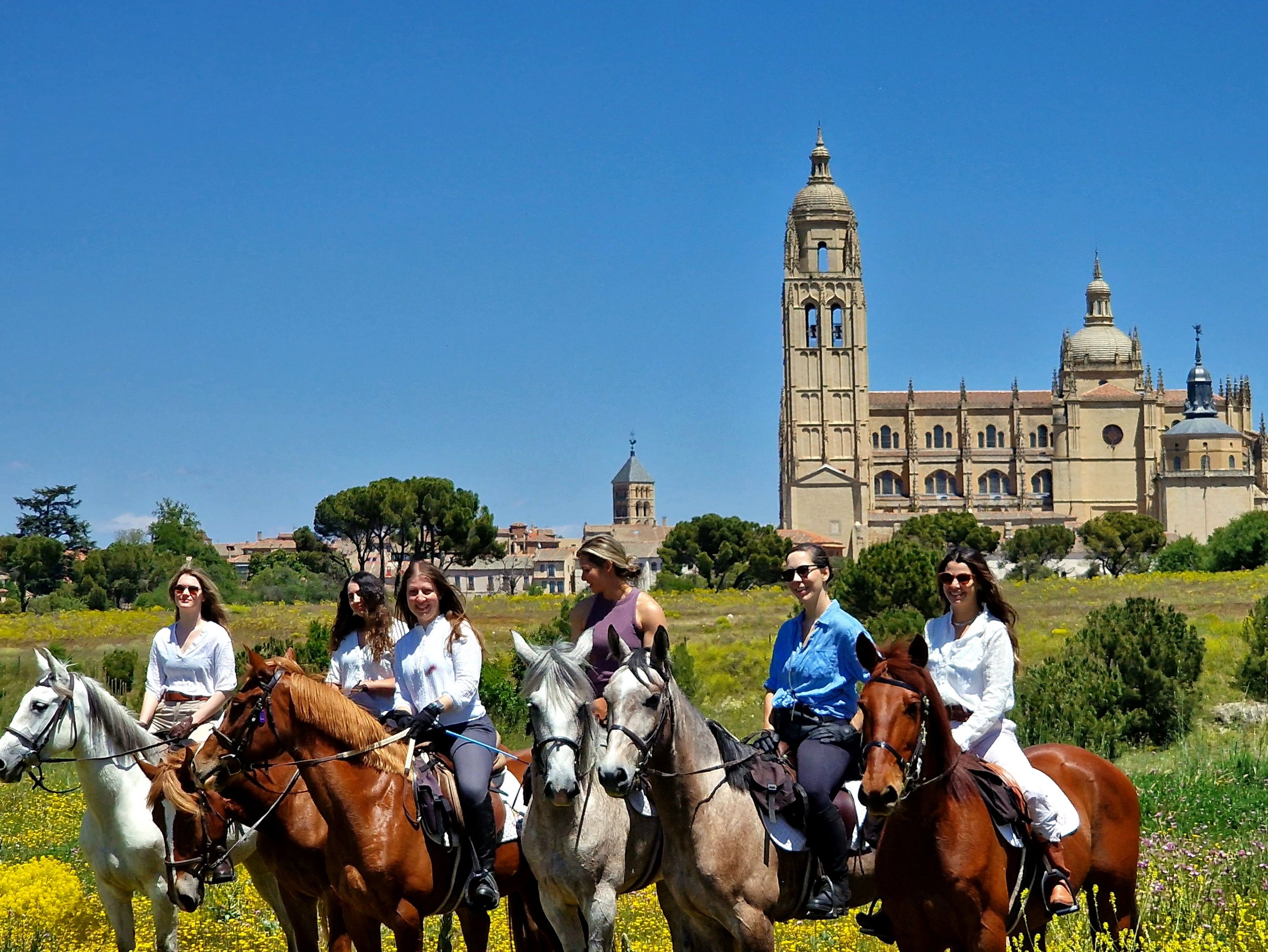 Six women riding horses in a field with a historic church and bright blue sky in the background.