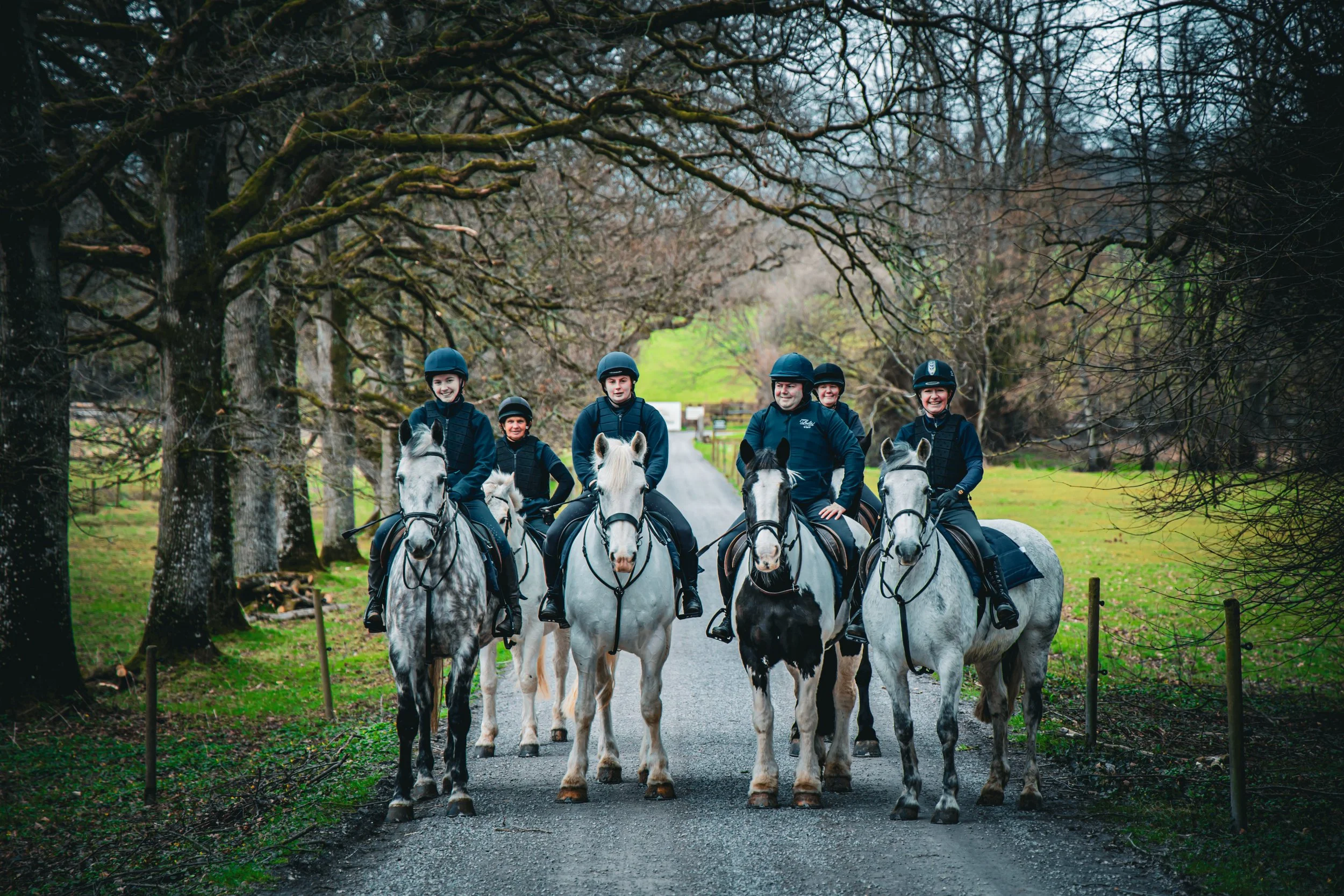 Five people riding horses on a gravel path through a wooded area, all wearing helmets and riding gear, with leafless trees and green grass in the background.