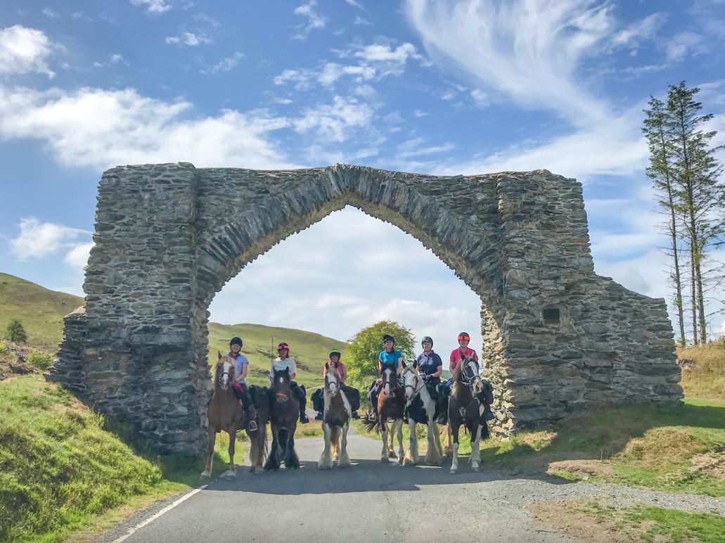 Six people riding horses under an ancient stone archway in a green countryside setting.