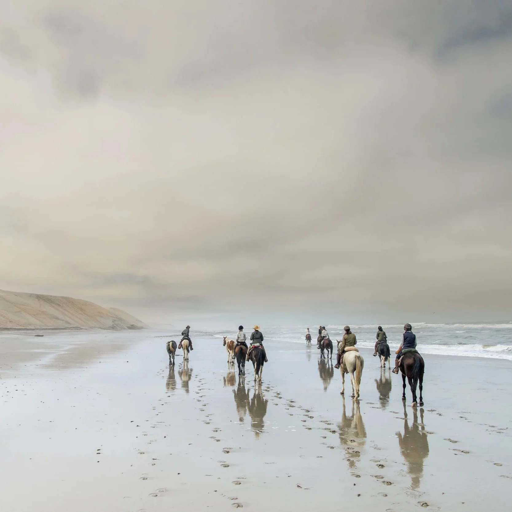 Group of people riding horses on a beach with cloudy sky and reflection on wet sand.