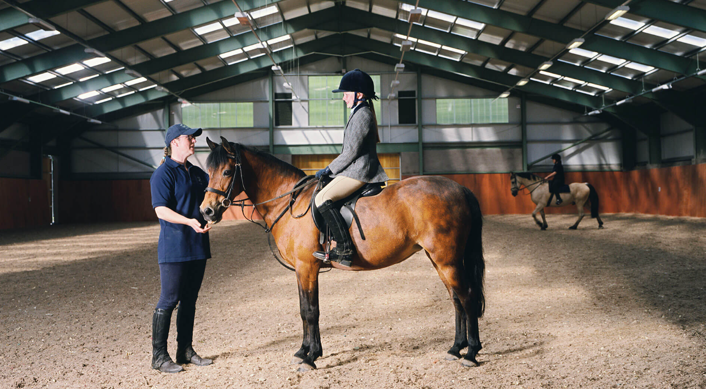 A woman riding a brown horse inside an indoor riding arena, speaking with a trainer who holds the horse's bridle. In the background, another rider on a white horse is practicing.