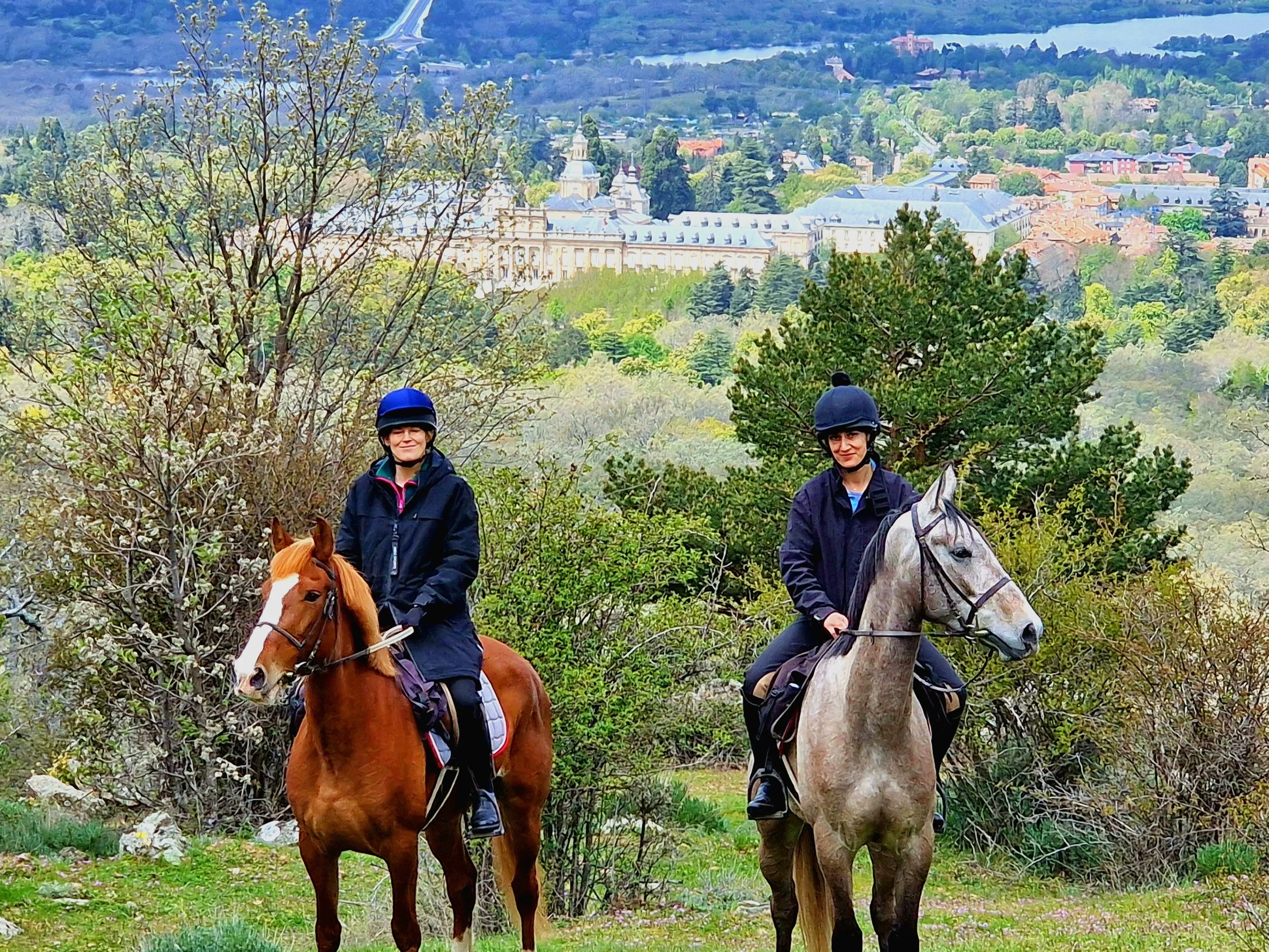 Two women riding horses in a lush green outdoor setting with a cityscape and historic buildings in the background.