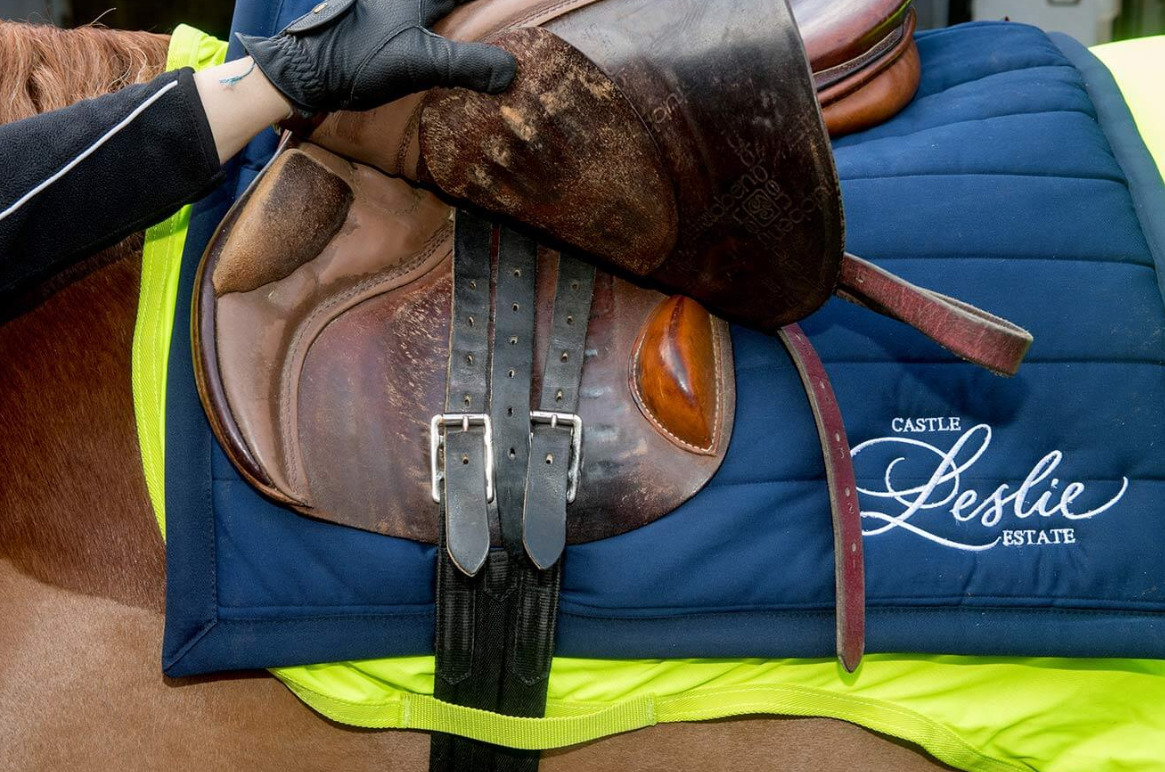 Close-up of a horse saddle placed on a blue saddle pad with 'Castle Leslie Estate' embroidered on it, on a brown horse, with a person's gloved hand adjusting the saddle.