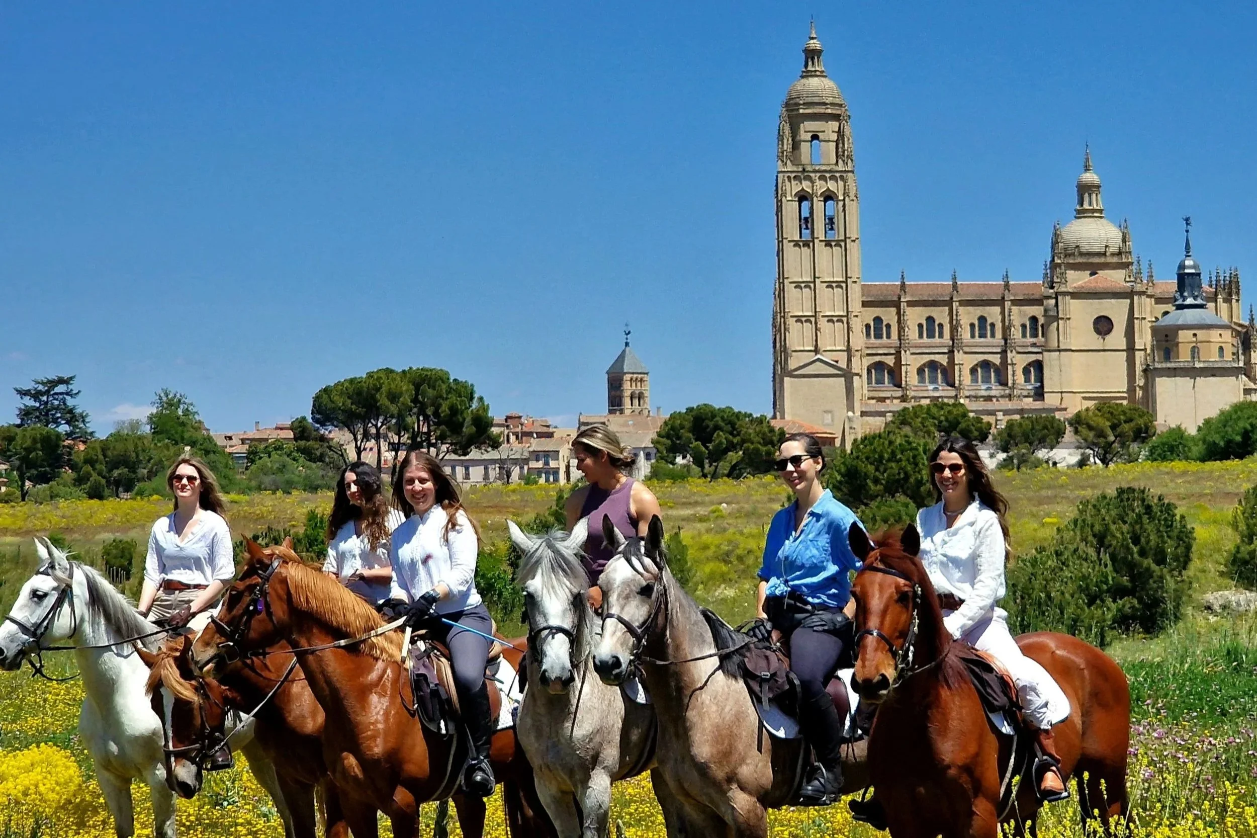 Six women riding horses through a grassy field with yellow flowers, with a large historic cathedral and trees in the background on a sunny day.