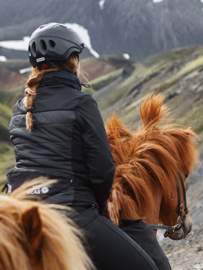 Person in black jacket and helmet riding a brown horse through mountainous terrain.