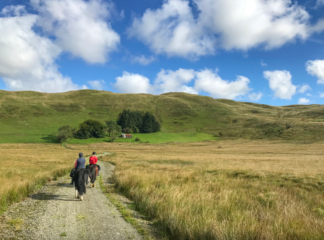 Two people riding horses down a dirt trail through a grassy field towards distant hills under a partly cloudy sky.