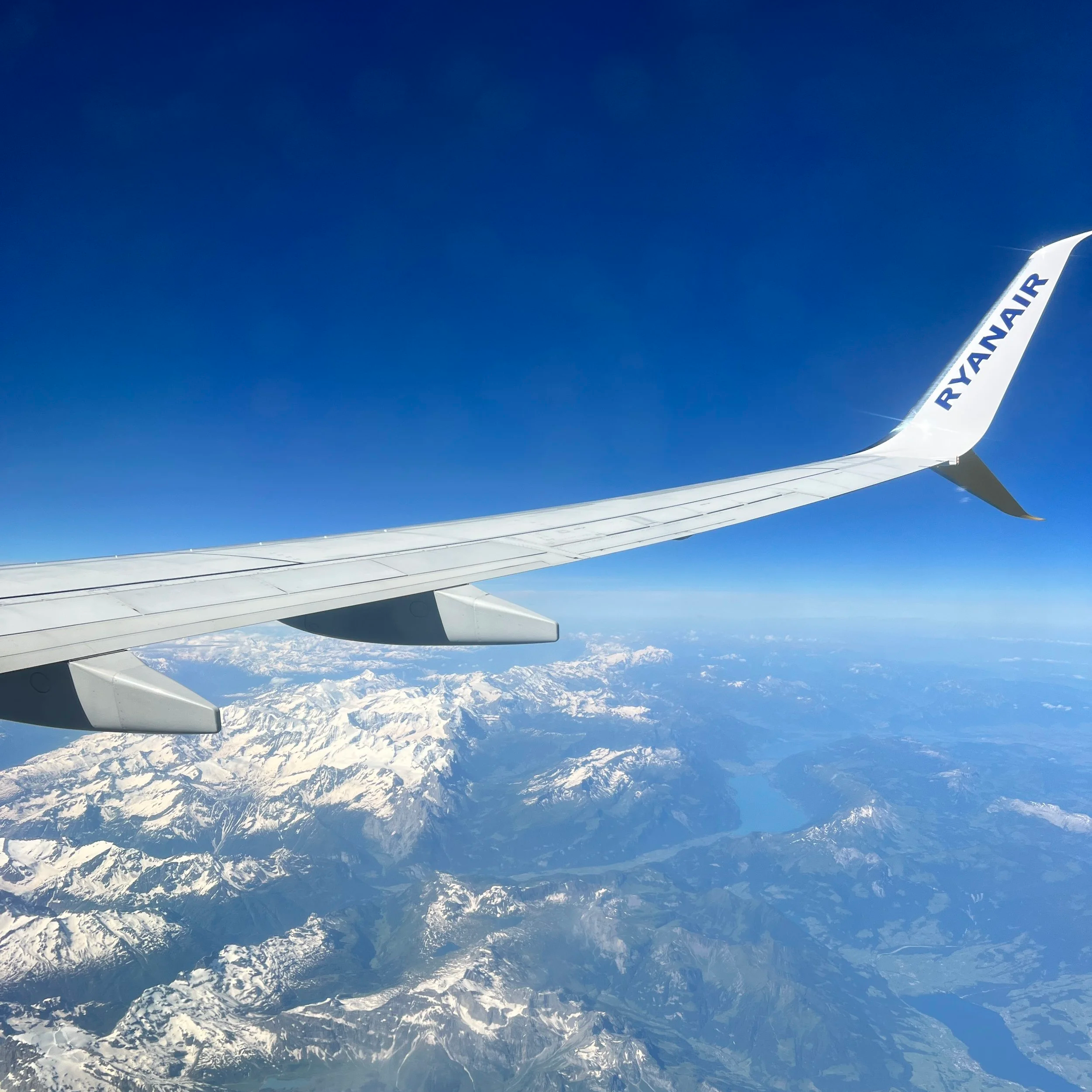 Photo taken from an airplane window showing the wing with 'RYANAIR' branding, snow-capped mountains, and a large lake below.