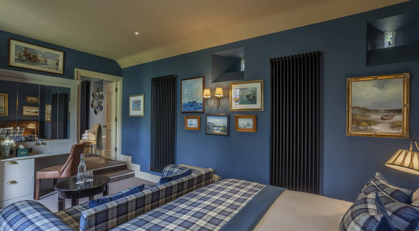 A cozy bedroom with blue walls decorated with framed nautical paintings and small windows. There is a plaid bed, a plaid sofa, a small round table with water bottles, and a mirror reflecting part of the room.