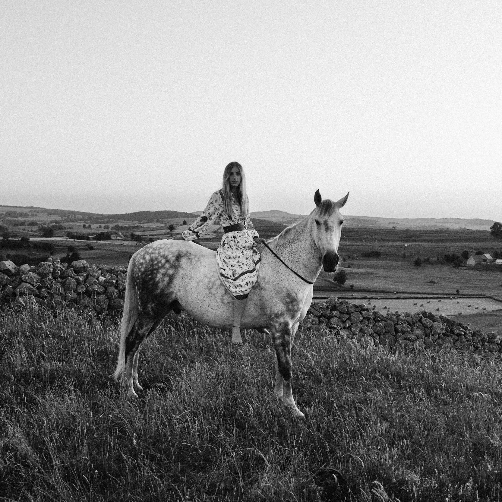 A woman riding a white horse with darker spots in a rural landscape with fields and hills in the background, black and white photo.