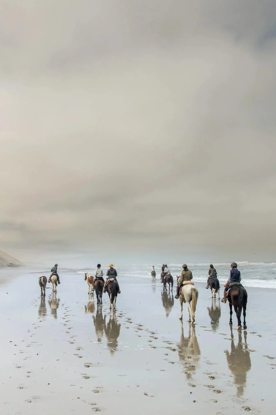 Group of people horseback riding on a beach with footprints in the sand and the ocean in the background.