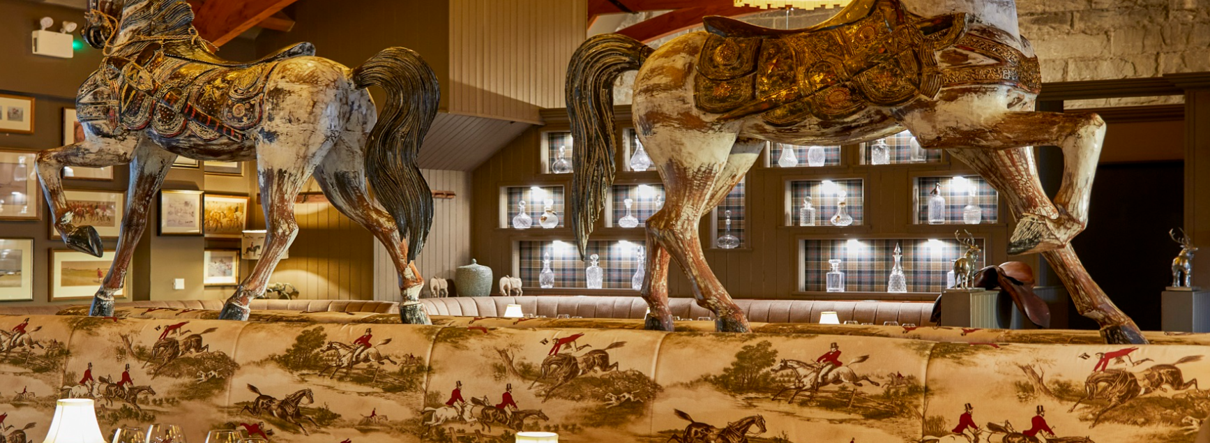 Interior of a room with two large wooden horse sculptures with intricate painted designs on their bodies, standing above a vintage-style upholstered sofa featuring a tapestry pattern of fox hunts.