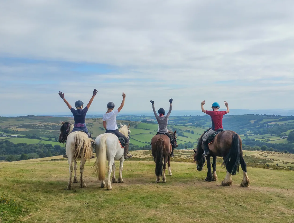 Four people riding horses on a hilltop with a scenic view of the countryside, waving their hands in the air.