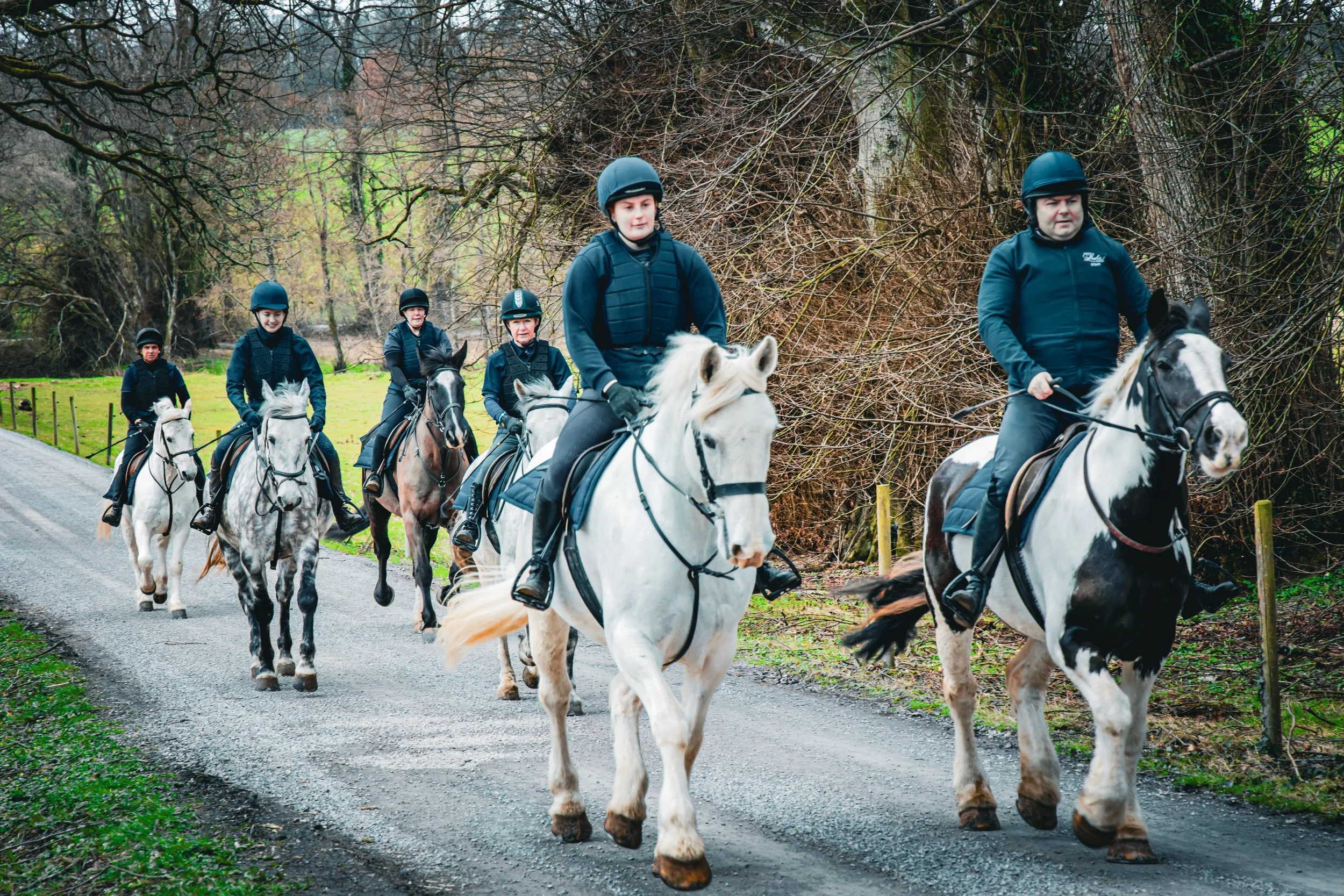 Group of six people riding horses on a gravel trail surrounded by leafless trees and greenery