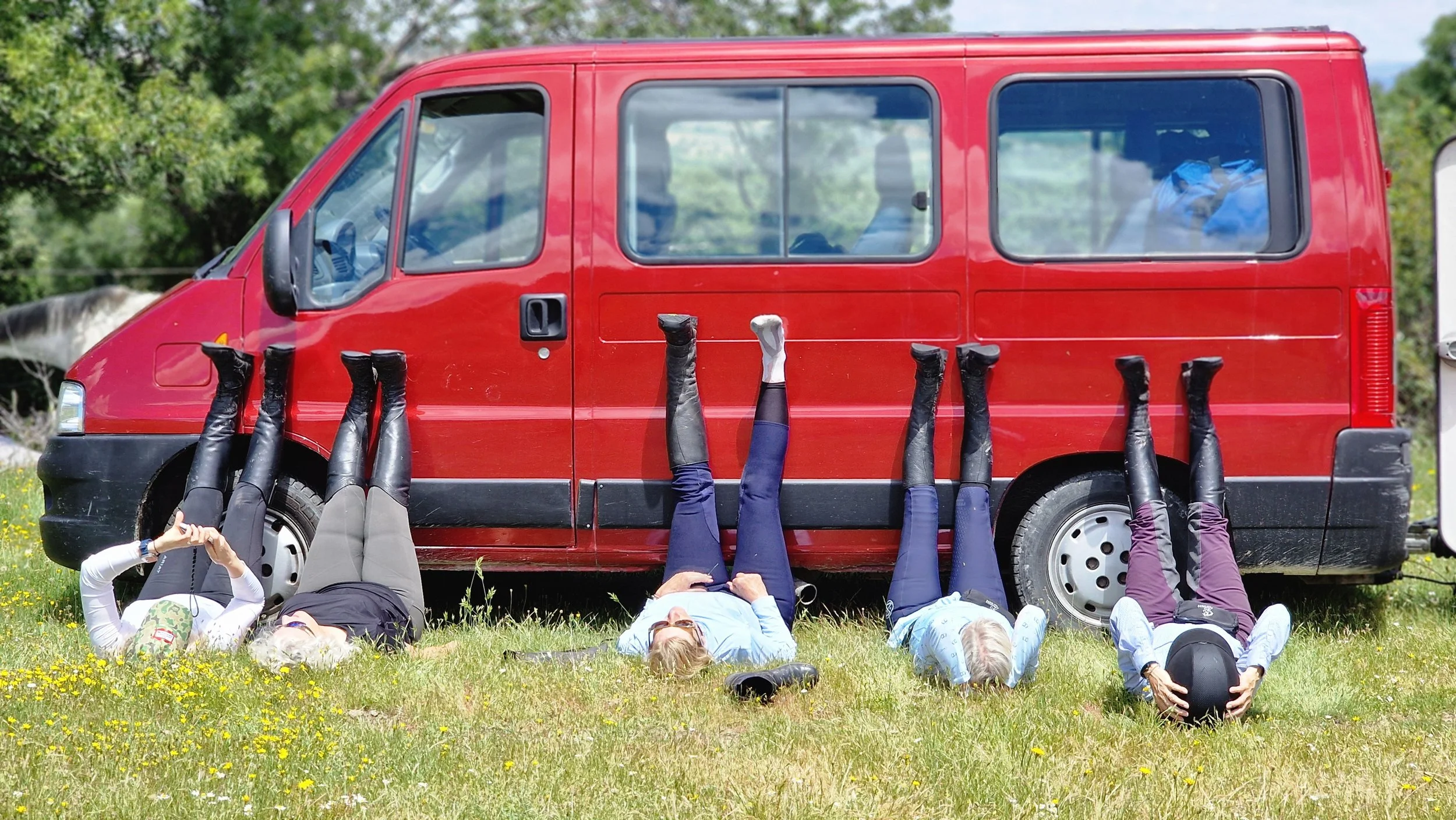 Four women lying on the grass with their legs up against a red van, outside in a grassy area with trees in the background.