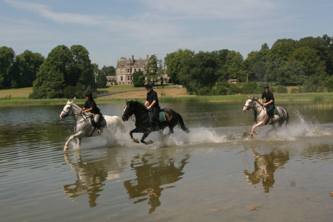 Three people riding horses through a shallow body of water with a historic mansion and trees in the background on a sunny day.