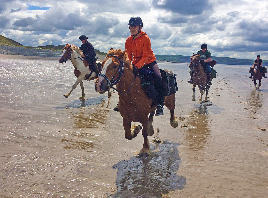 People horseback riding along the shoreline of a beach with cloudy skies overhead.