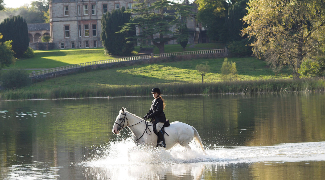 A woman riding a white horse through a body of water with a castle or large house, green trees, and a grassy hill in the background.