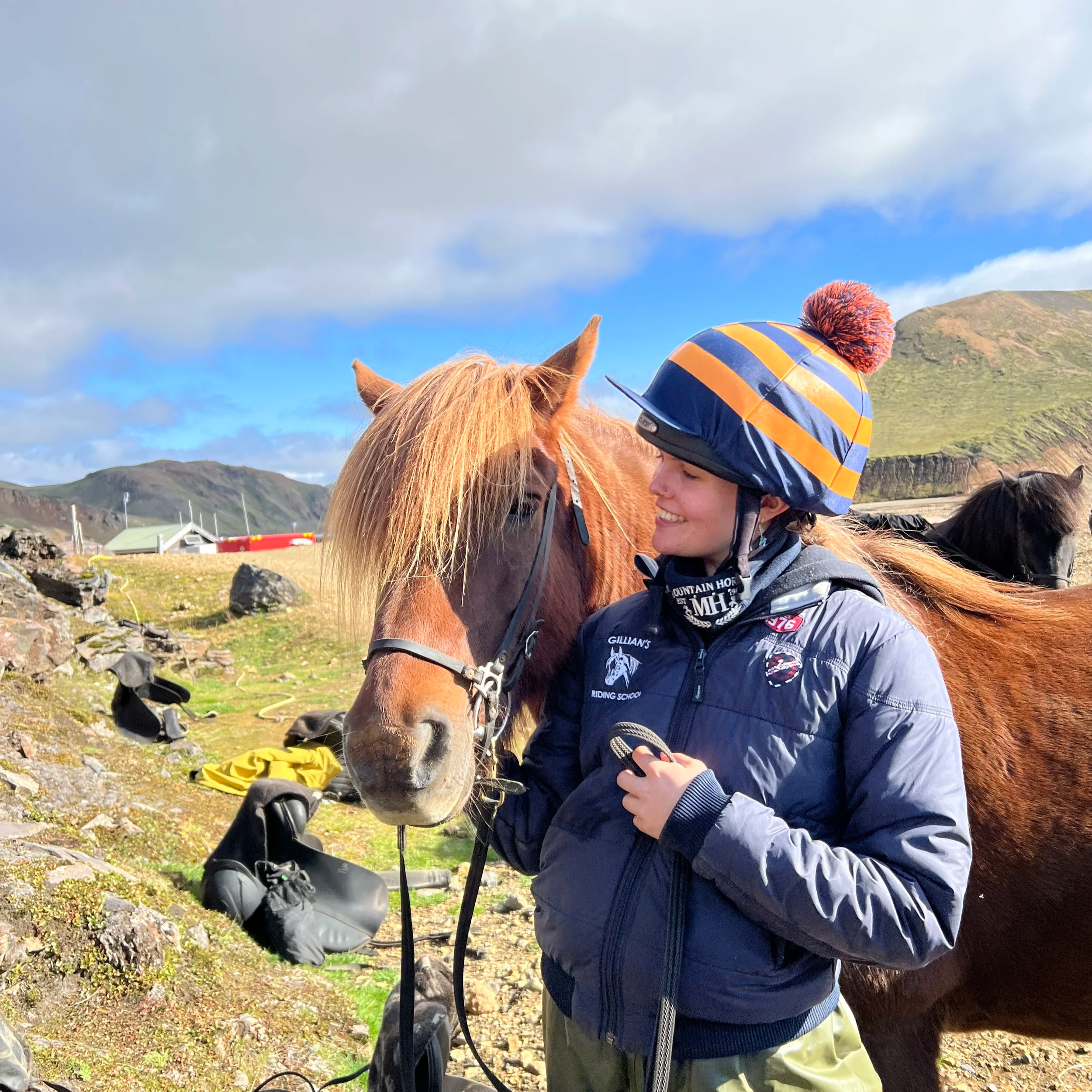 A woman smiling and holding a phone while standing next to a brown horse outdoors in a mountainous area with blue sky and clouds.