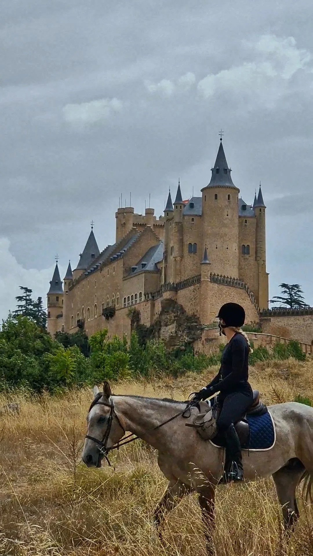 A woman riding a horse in front of a large castle with towers and stone walls, under a cloudy sky.