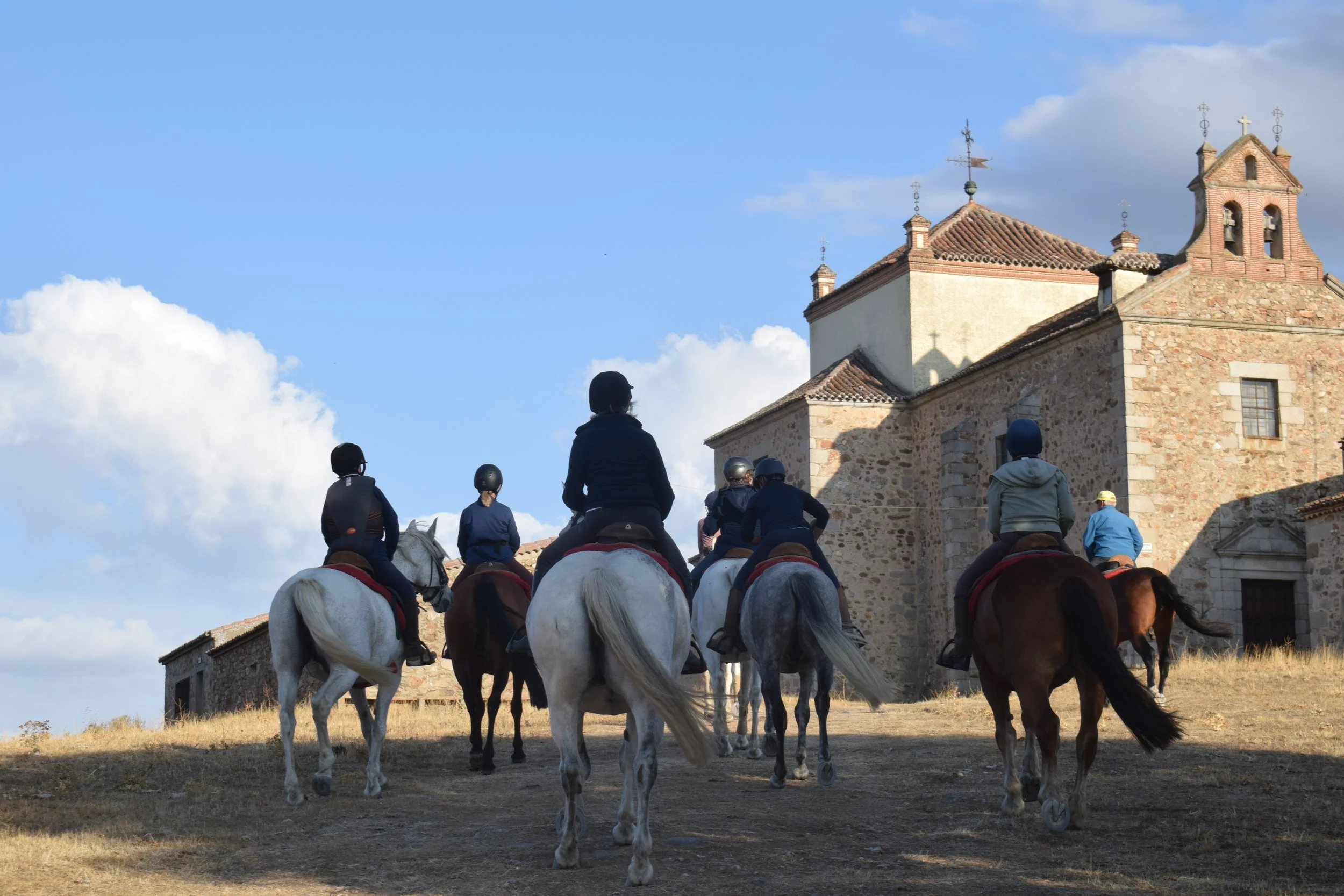Five people riding horses and wearing helmets near an old stone church under a partly cloudy sky.