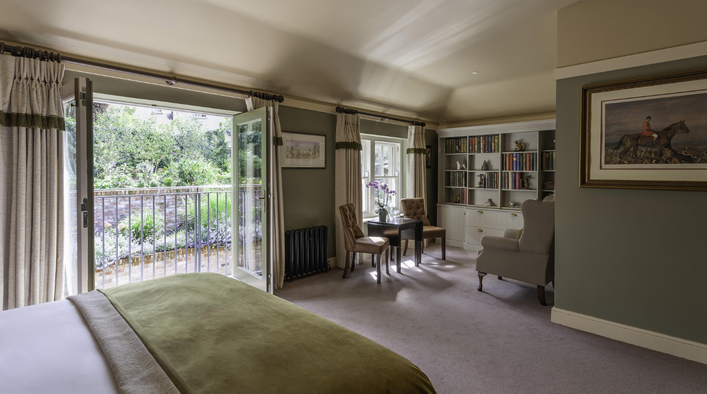 A bedroom with an open door leading to a balcony garden, a bed with green bedding, a sitting area with two chairs and a small table, and built-in bookshelves filled with books and decorative items.
