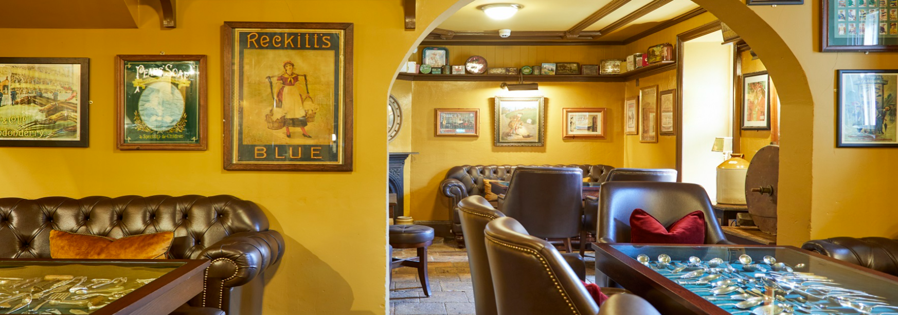 Interior of a cozy vintage-style pub with yellow walls, leather tufted armchairs, framed artwork, and a display of silverware on a table, with a doorway leading to another seating area.