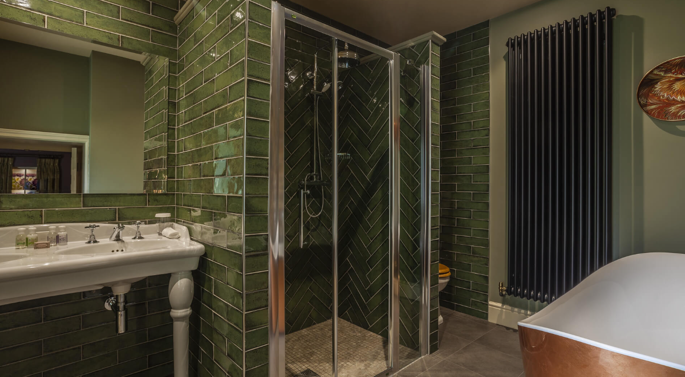 Bathroom with green tiled walls, a shower stall with glass door, a white pedestal sink with toiletries, a large black radiator, and a white bathtub with wood paneling.