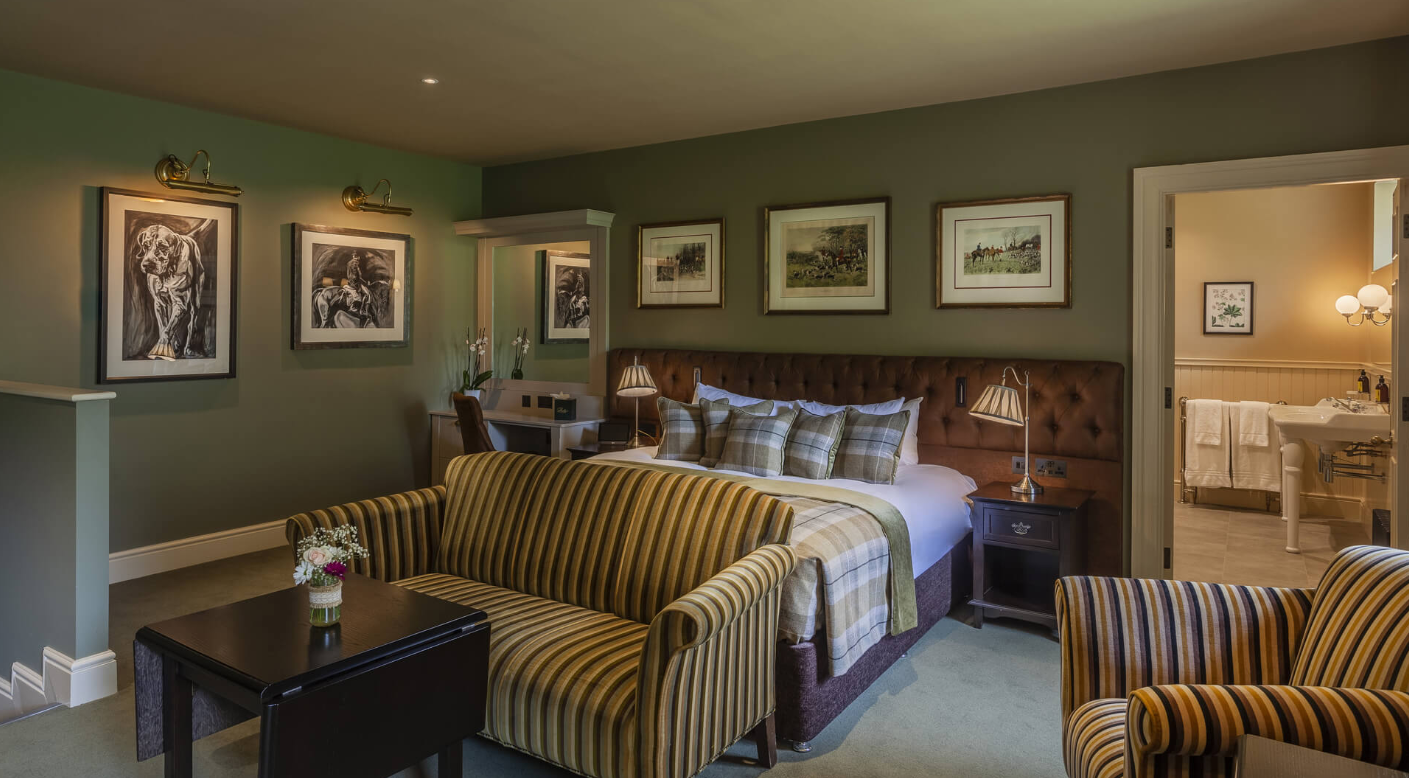 A cozy hotel room with a large bed, striped armchair, striped sofa, bedside tables with lamps, framed artwork on the walls, and a view into a bathroom with towels and a sink.