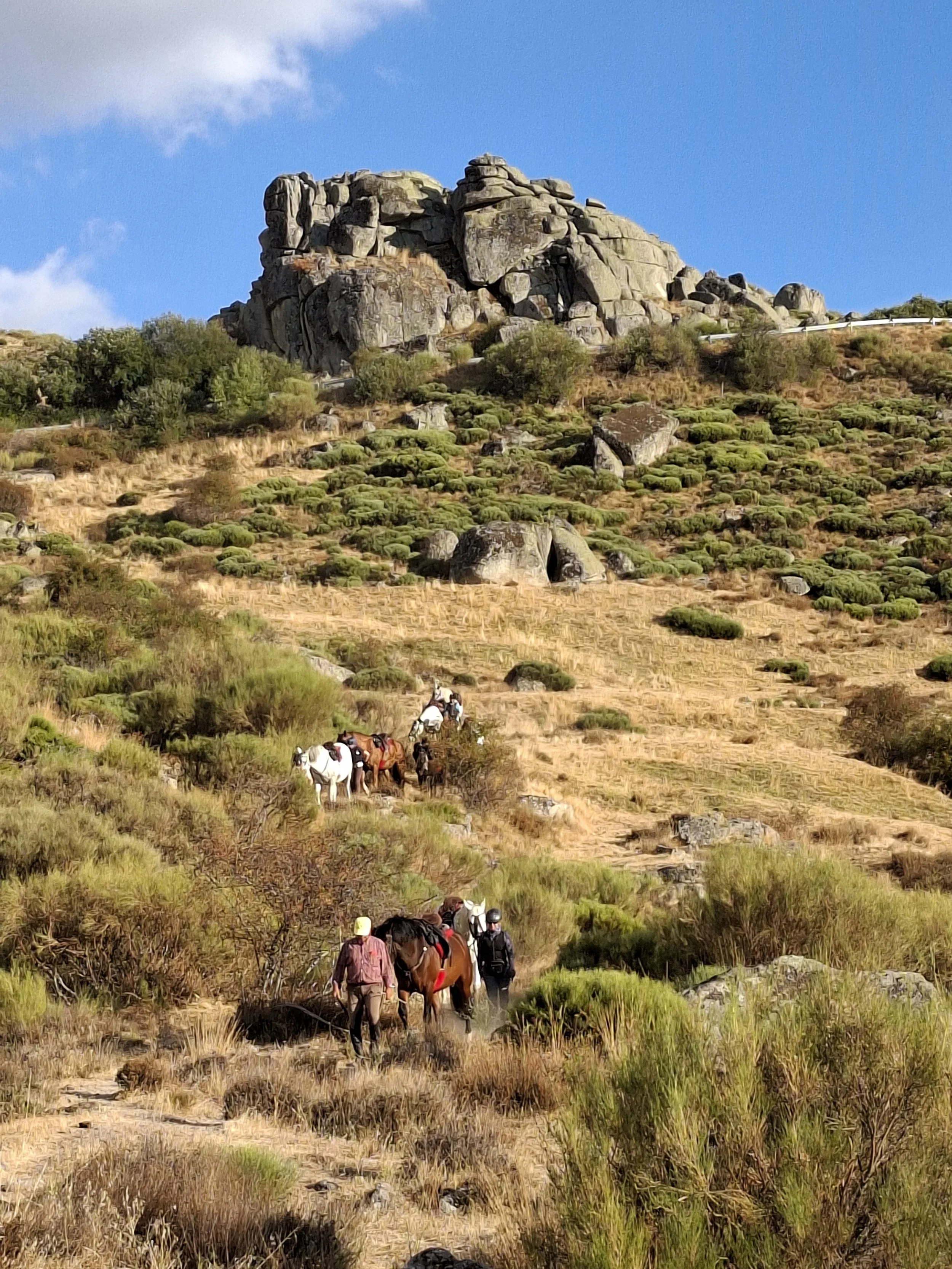 Horse riding holiday in Spain with horses walking on a trail in a mountainous landscape with rocks, bushes, and a partly cloudy blue sky.