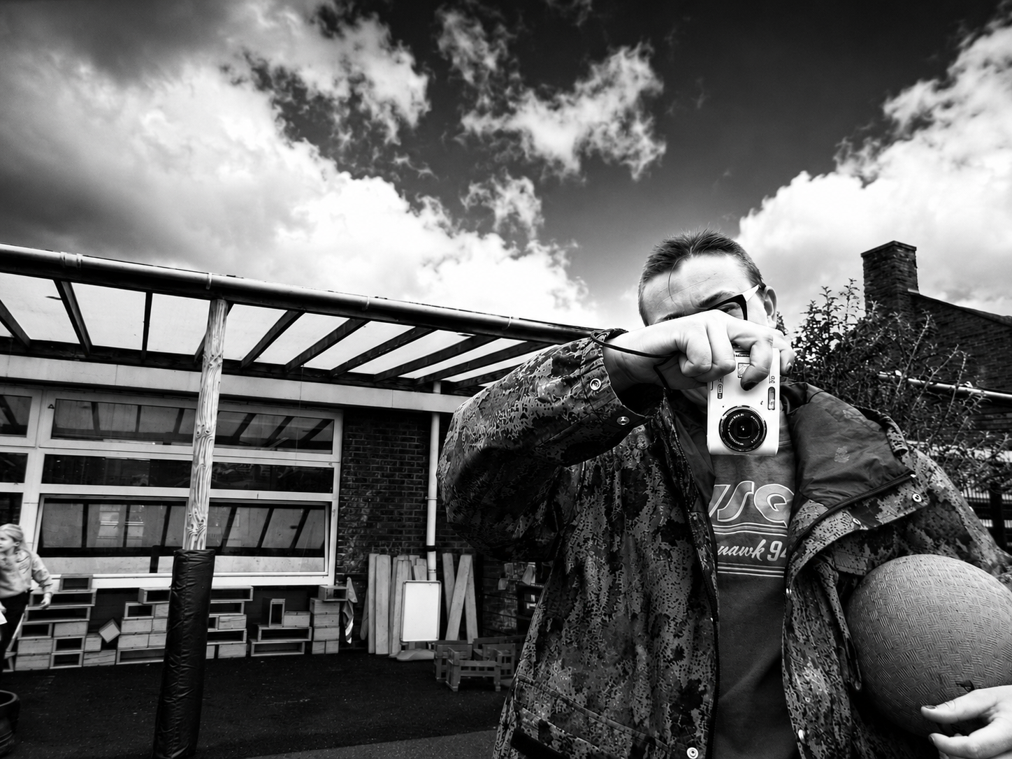 A person holding a camera up to take a picture outdoors, with a building and children in the background against a cloudy sky, in black and white. Photo Explorers After School Club London