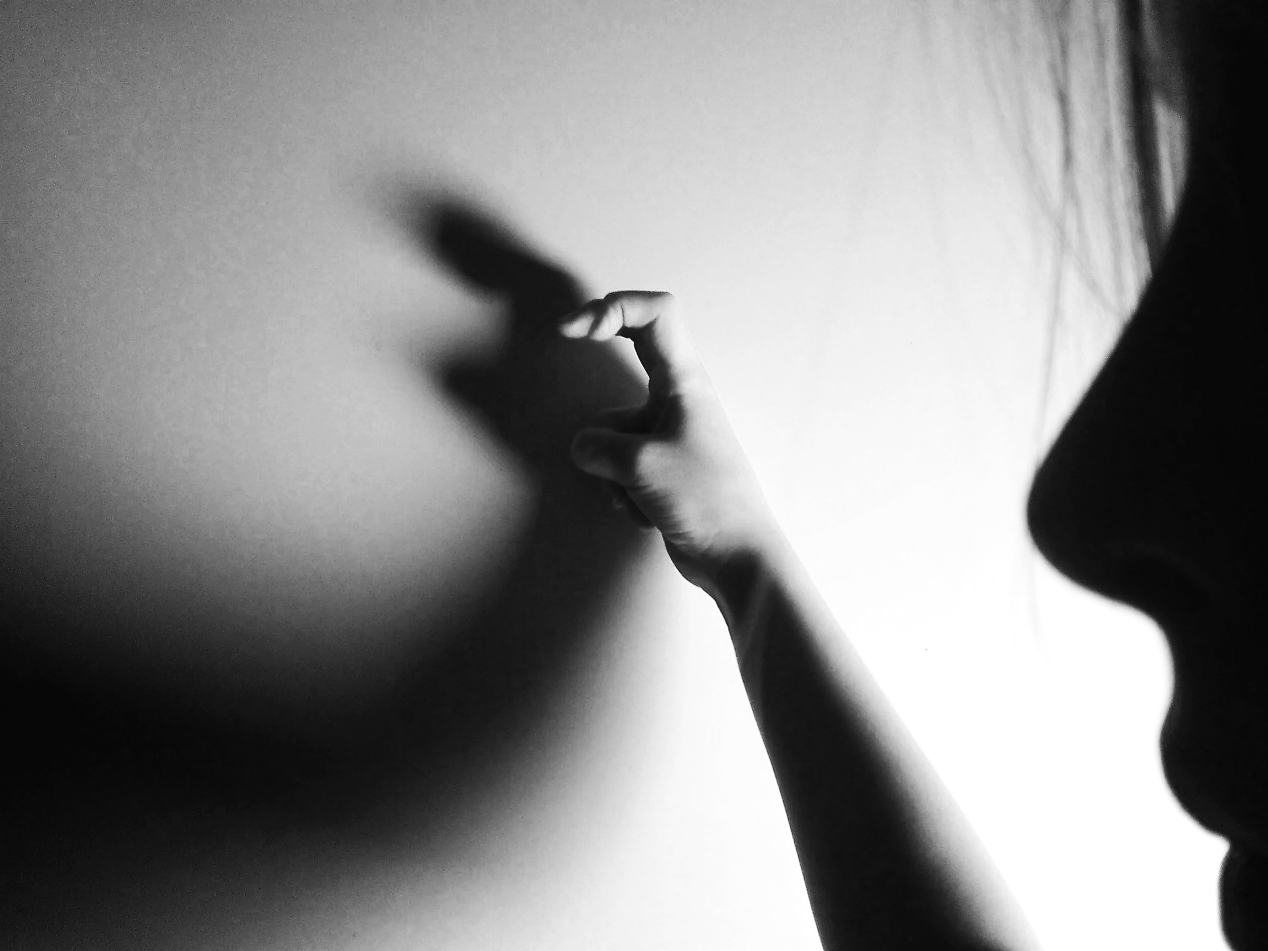 A black-and-white side profile of a person reaching out and touching a wall with their finger, casting a shadow of their hand on the wall.