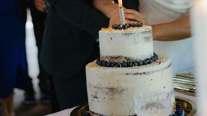 Two-tiered white wedding cake decorated with blueberries, with a lit candle on top, being cut by a couple at a celebration.