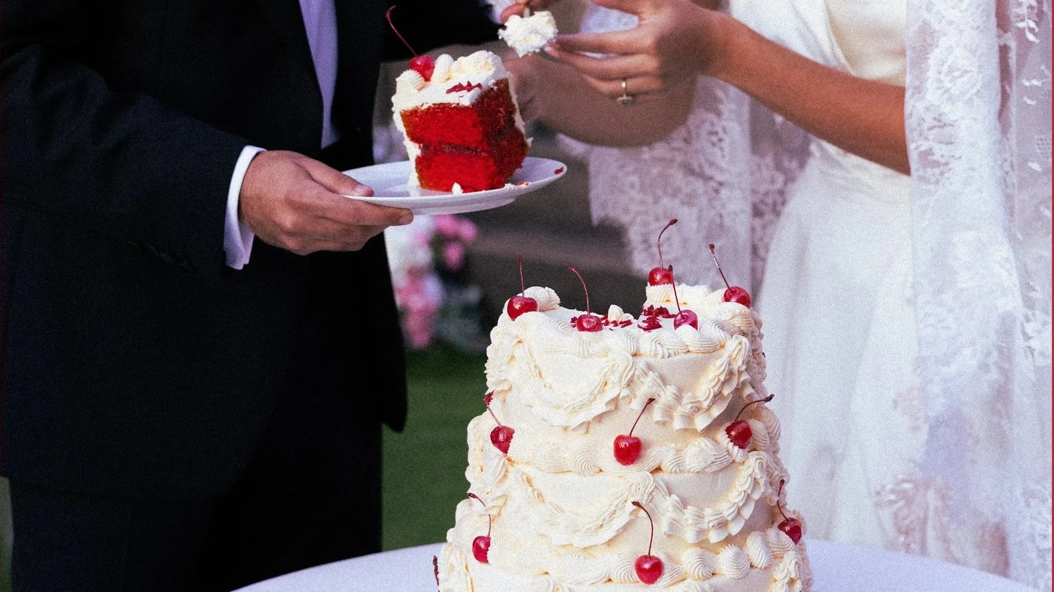 Wedding cake with cherries on top and a person in a suit holding a plate with a slice of cake.