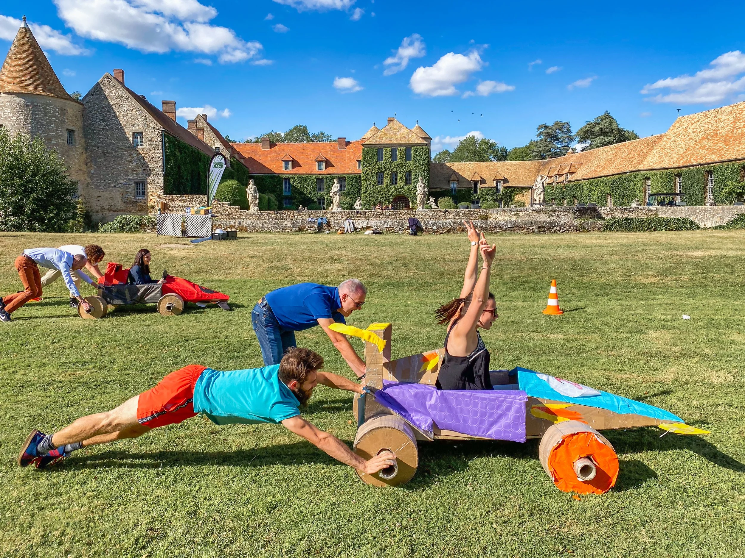 Groupe de personnes participant à une course de voitures en carton dans un grand espace vert, avec un vieux château en arrière-plan sous un ciel bleu avec quelques nuages.