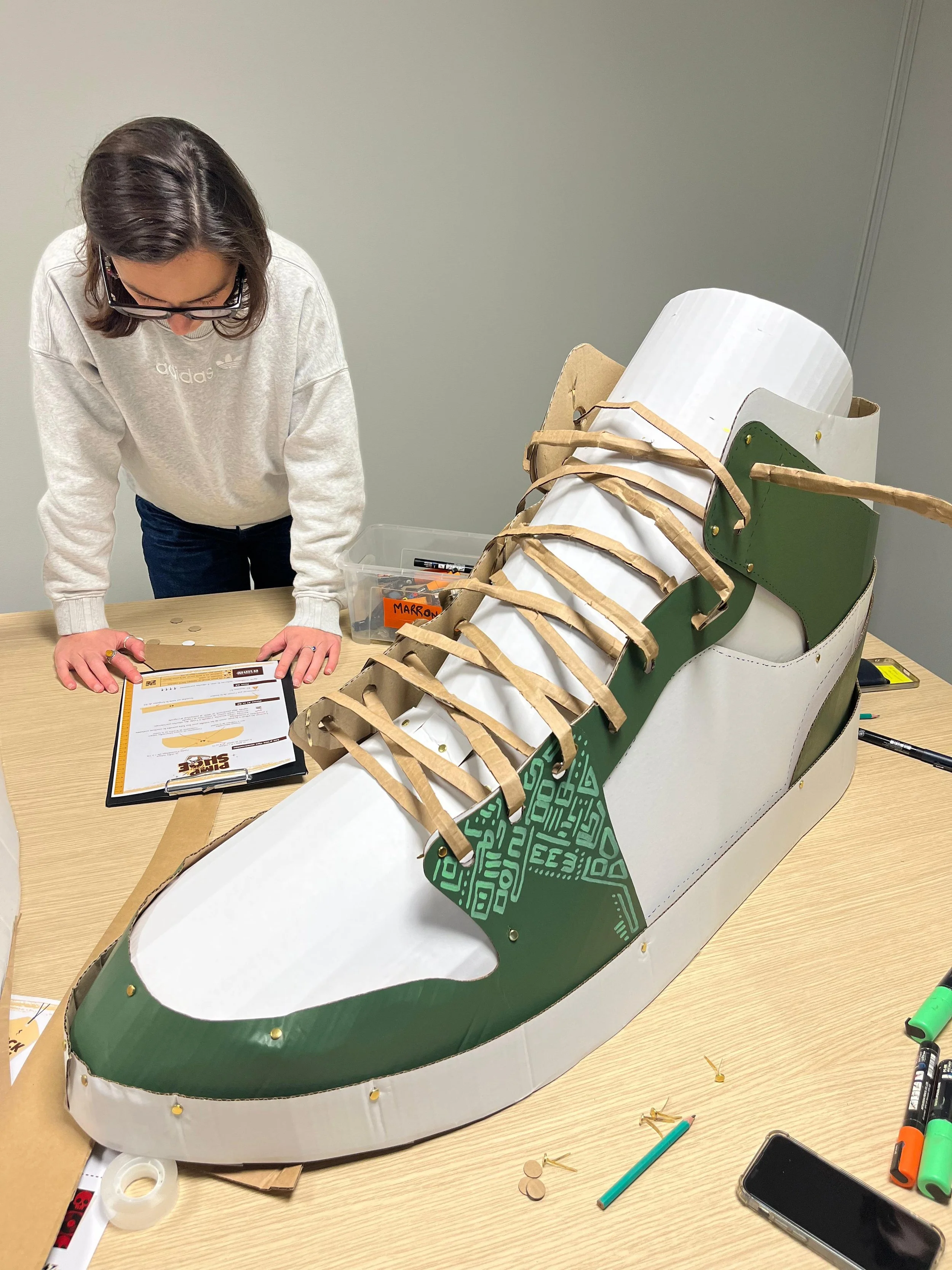 Chaussure en carton immense sur une table avec une femme qui réfléchie à sa création