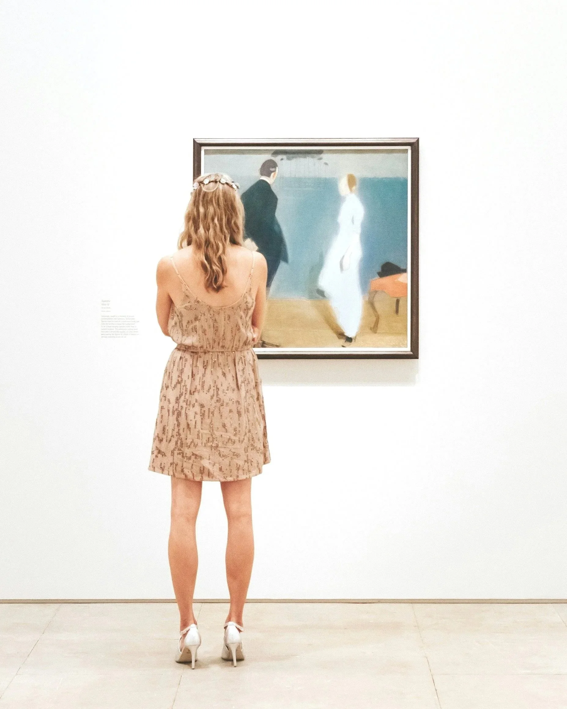 A woman with long, wavy hair wearing a beige patterned dress and white high heels stands in front of a framed painting in an art gallery, viewing the artwork.