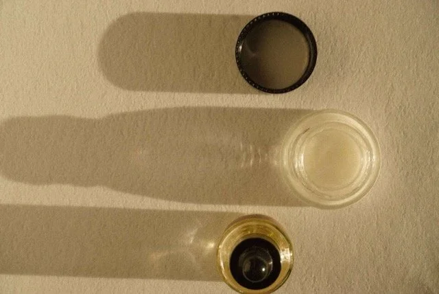 Top-down view of a light-colored kitchen countertop with a black cup filled with a light-colored beverage, a clear glass of water, and a small container with a black lid. Shadows cast by the objects are visible.