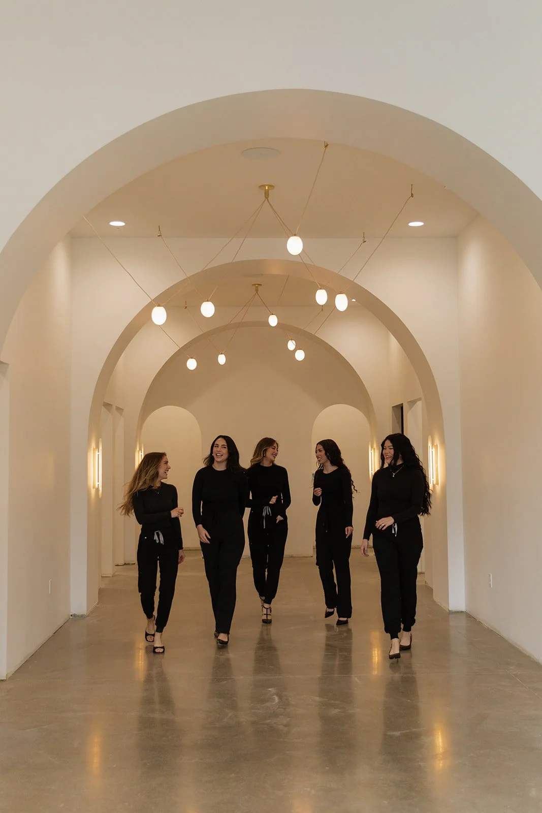 Five women wearing black outfits and high heels walking in a hallway with white walls and arched ceilings, illuminated by hanging globe pendant lights.