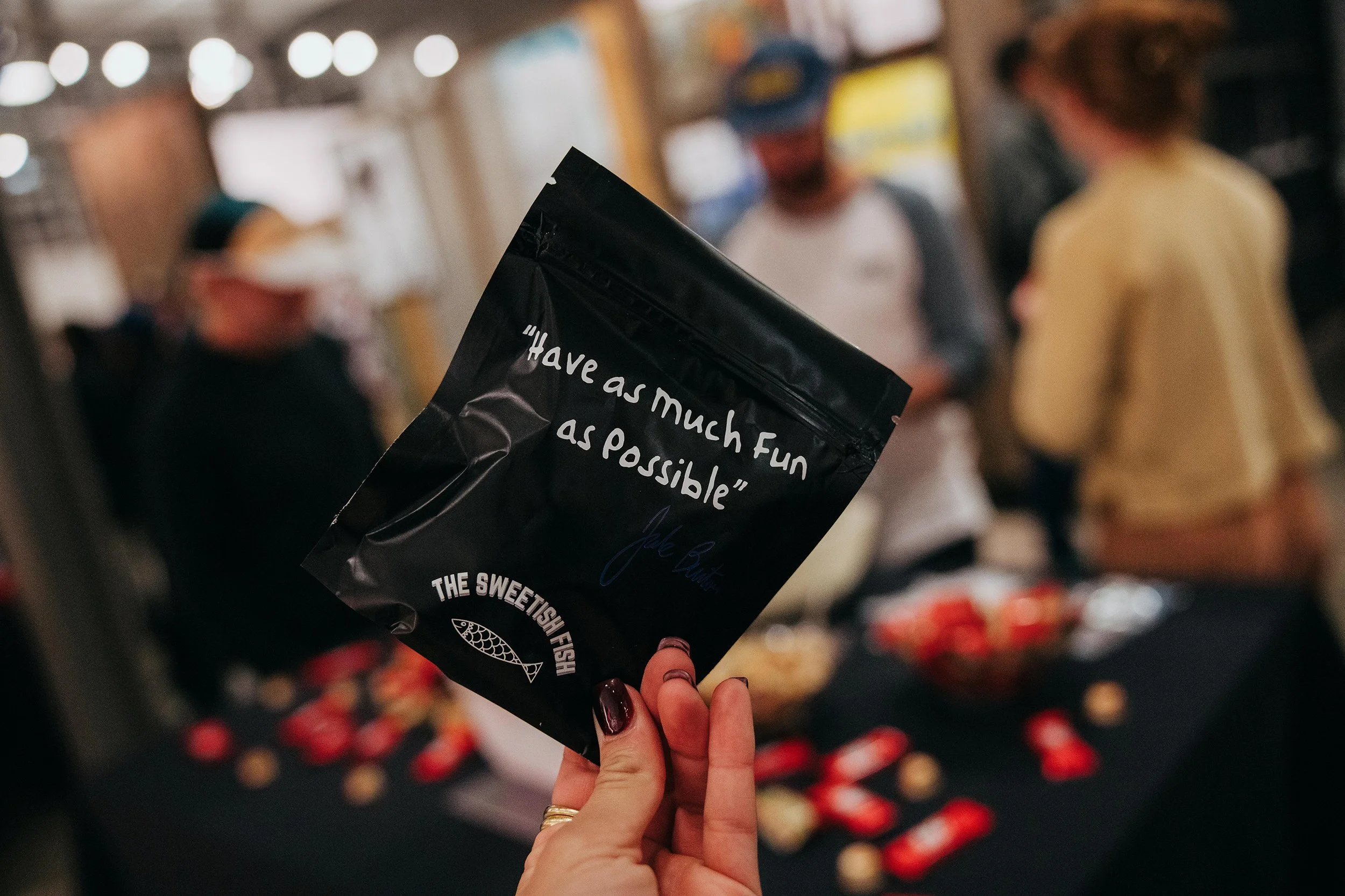 Hand holding a black bag with the quote 'Have as much fun as possible' at a social gathering with people in the background.