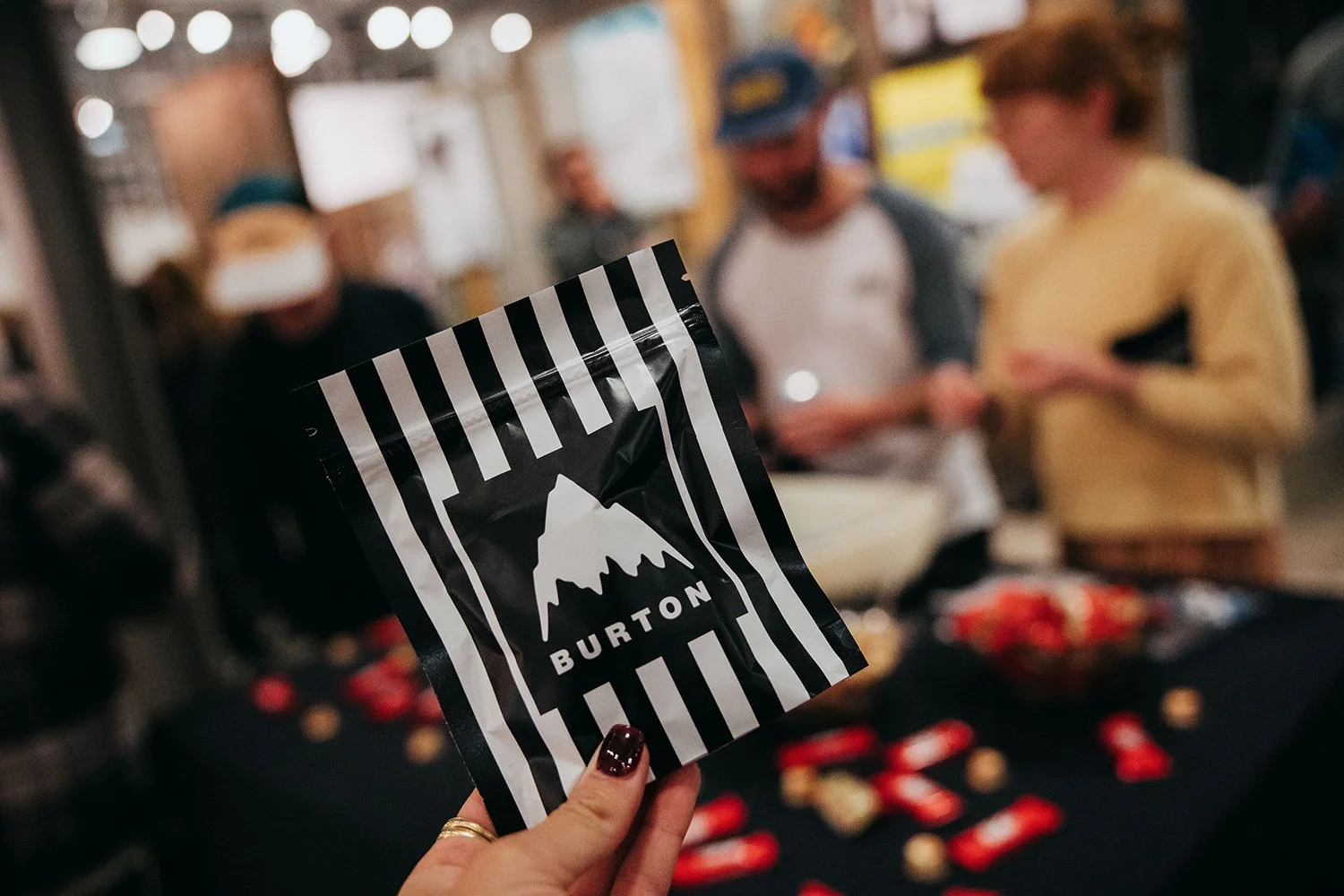 Close-up of a person holding a black and white striped packet with a mountain logo and the word "Burton" in front of a blurred group of people at a gathering or party.