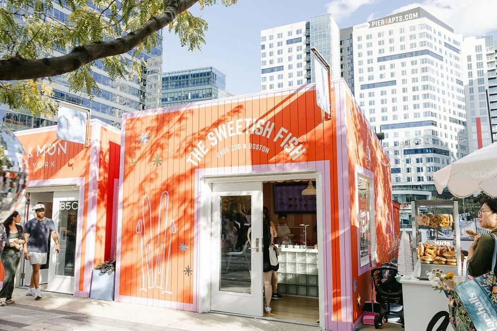Bright orange food stand called "The Sweetfish Fish" with a white outline of a fish with two antennae. There are people outside the stand and a woman placing an order. Tall buildings and a clear sky are in the background.
