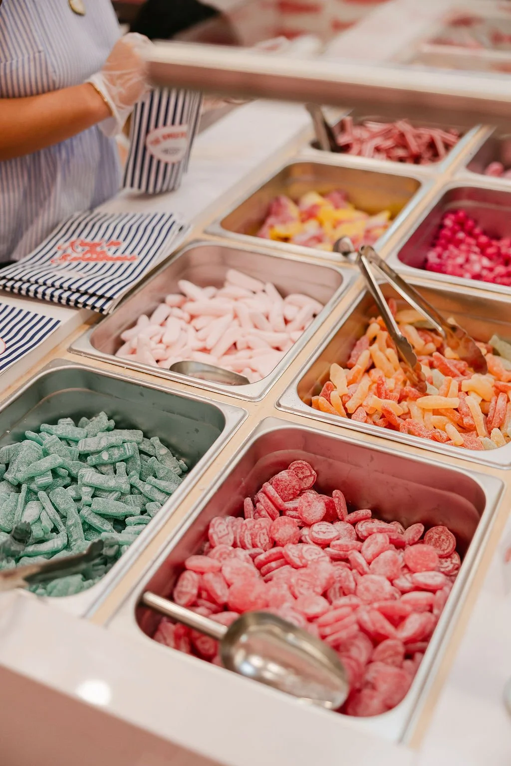 A variety of colorful Swedish and gummy candies in metal containers at an ice cream or candy shop, including green sour candies and red heart-shaped candies.