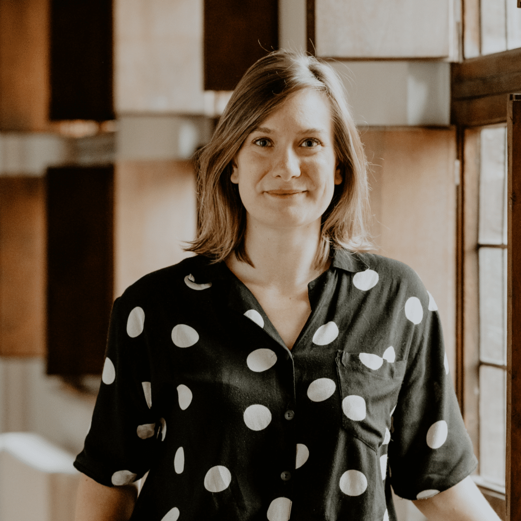 A young woman with shoulder-length light brown hair, wearing a black shirt with white polka dots, standing indoors near wooden-framed windows.
