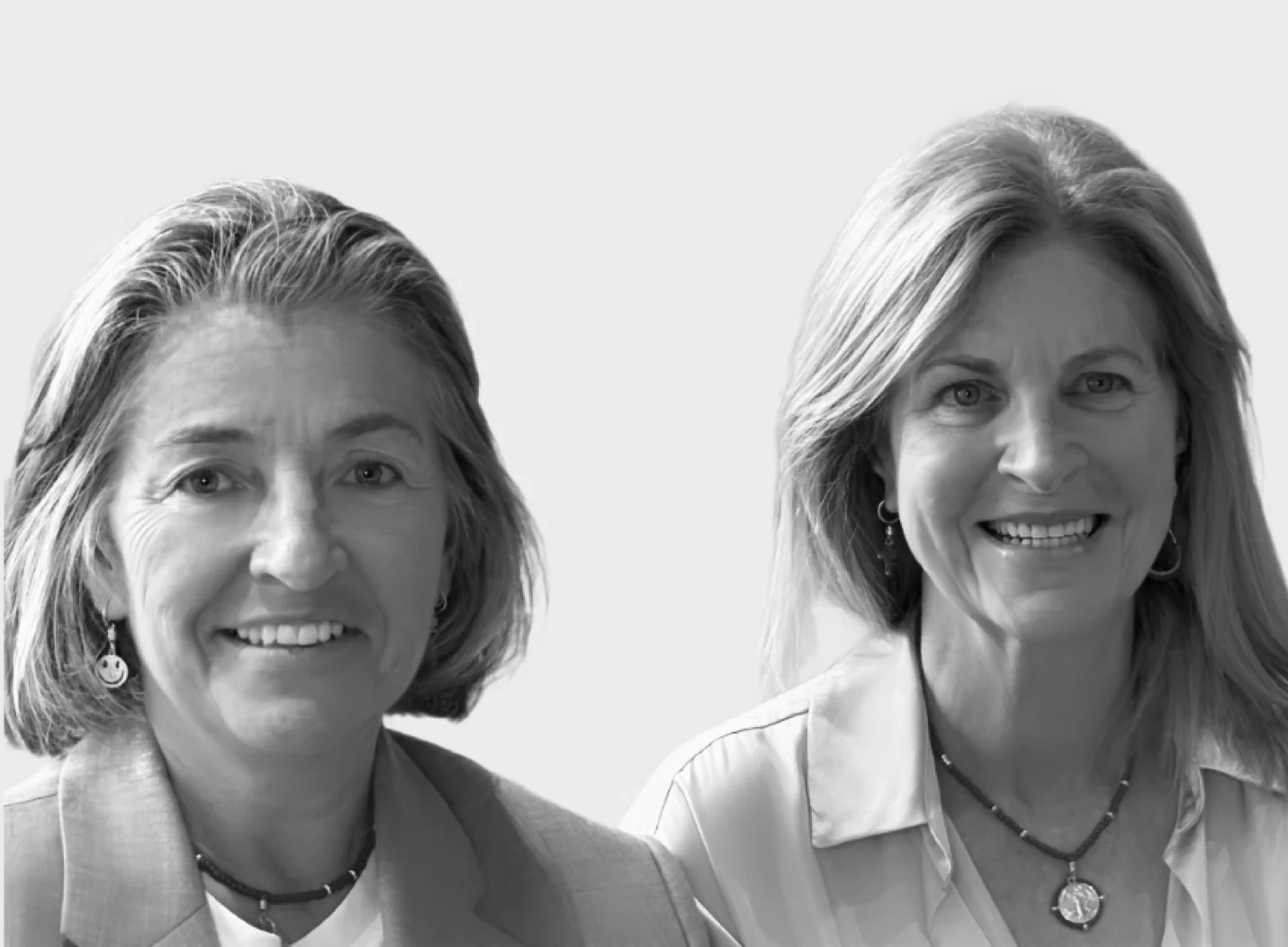 Black and white photo of two women smiling, with shoulder-length hair, wearing jewelry, and professional attire, against a plain background.