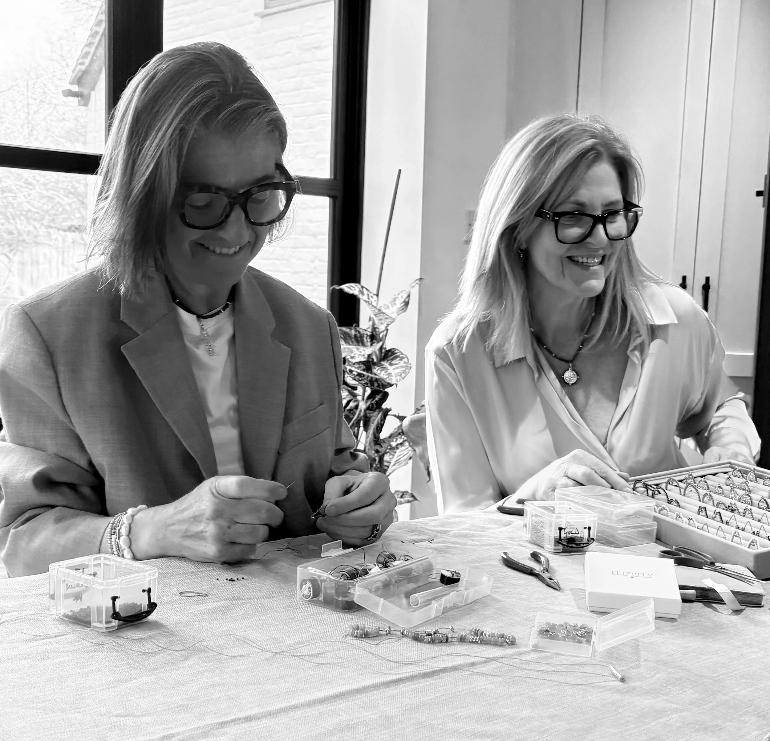 Two women sitting at a table, smiling, and working on jewelry or small craft items. There are small containers with jewelry pieces, tools, and jewelry displays on the table.
