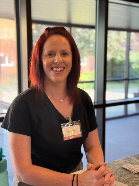 Smiling woman with red hair wearing a black shirt and name badge, seated at a table in a modern indoor setting with large windows and outdoor scenery visible in the background.