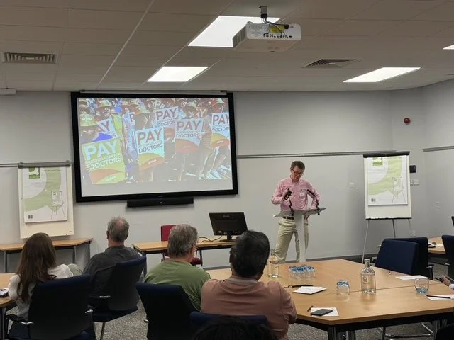 A man giving a presentation in a conference room with a large screen showing a protest with people holding signs that say 'PAY DOCTORS' or similar messages.